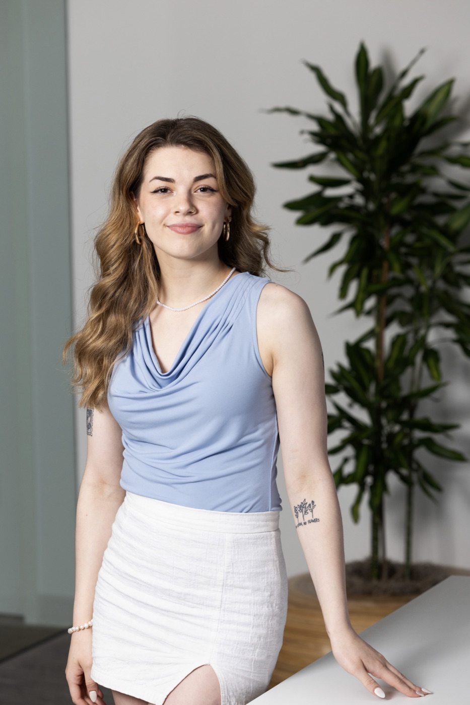 Emily Weinberg standing beside a tall potted plant in a bright room, in a pale blue top and white skirt, smiling at the camera.