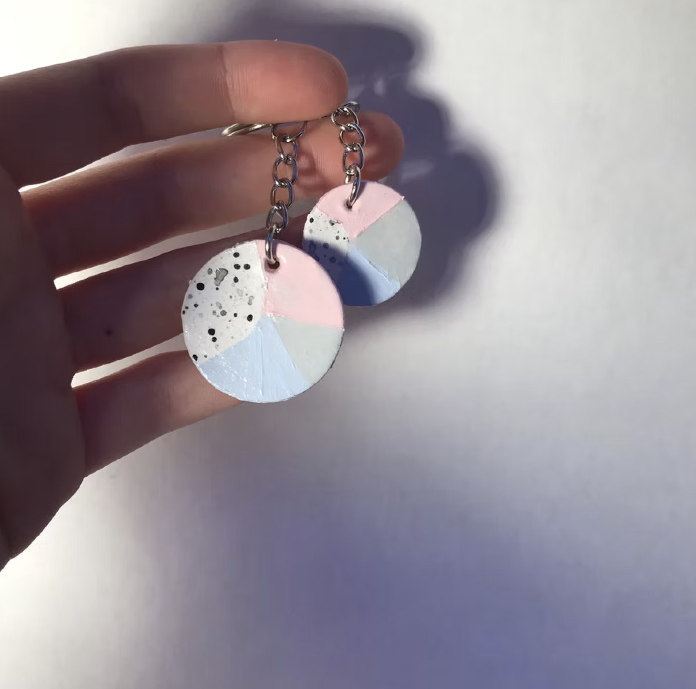 A pair of round polymer clay earrings held in one hand, quartered in pink, pale blue and white with gold flecks.