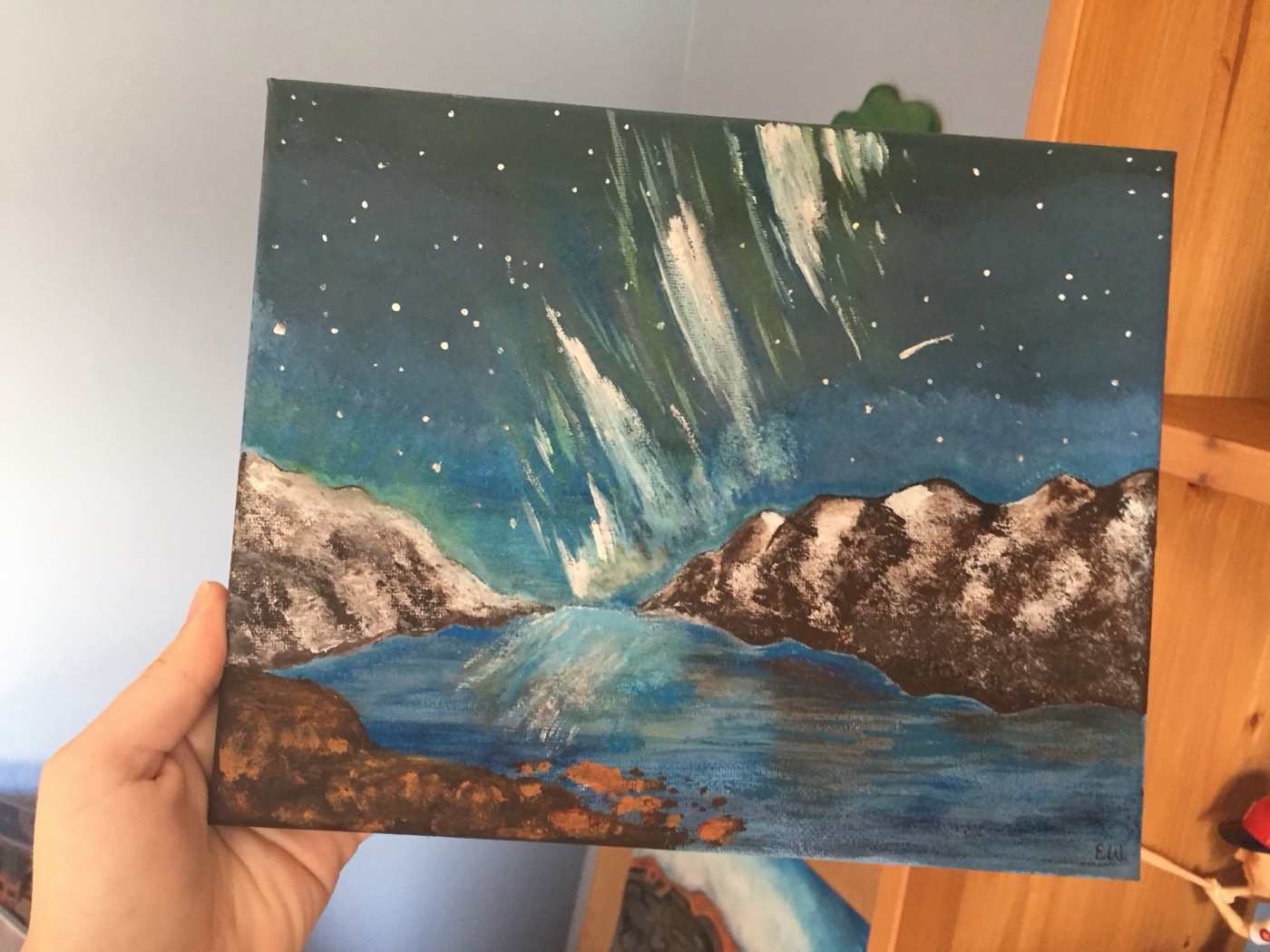 An acrylic painting of green and white northern lights over snow-topped mountains and dark water, held up in one hand.