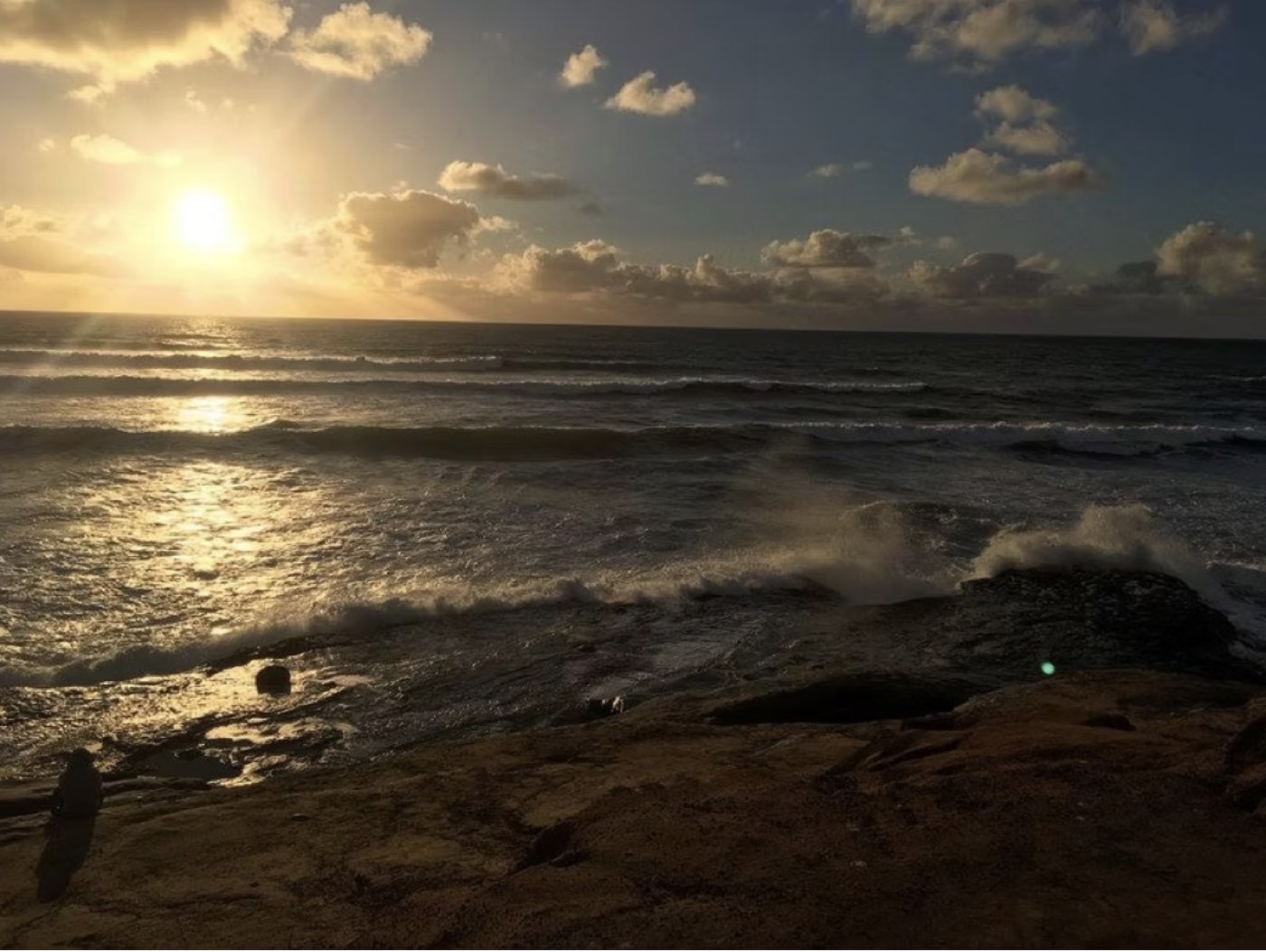 Low sun over the ocean, seen from a rock shelf as a wave breaks against it.